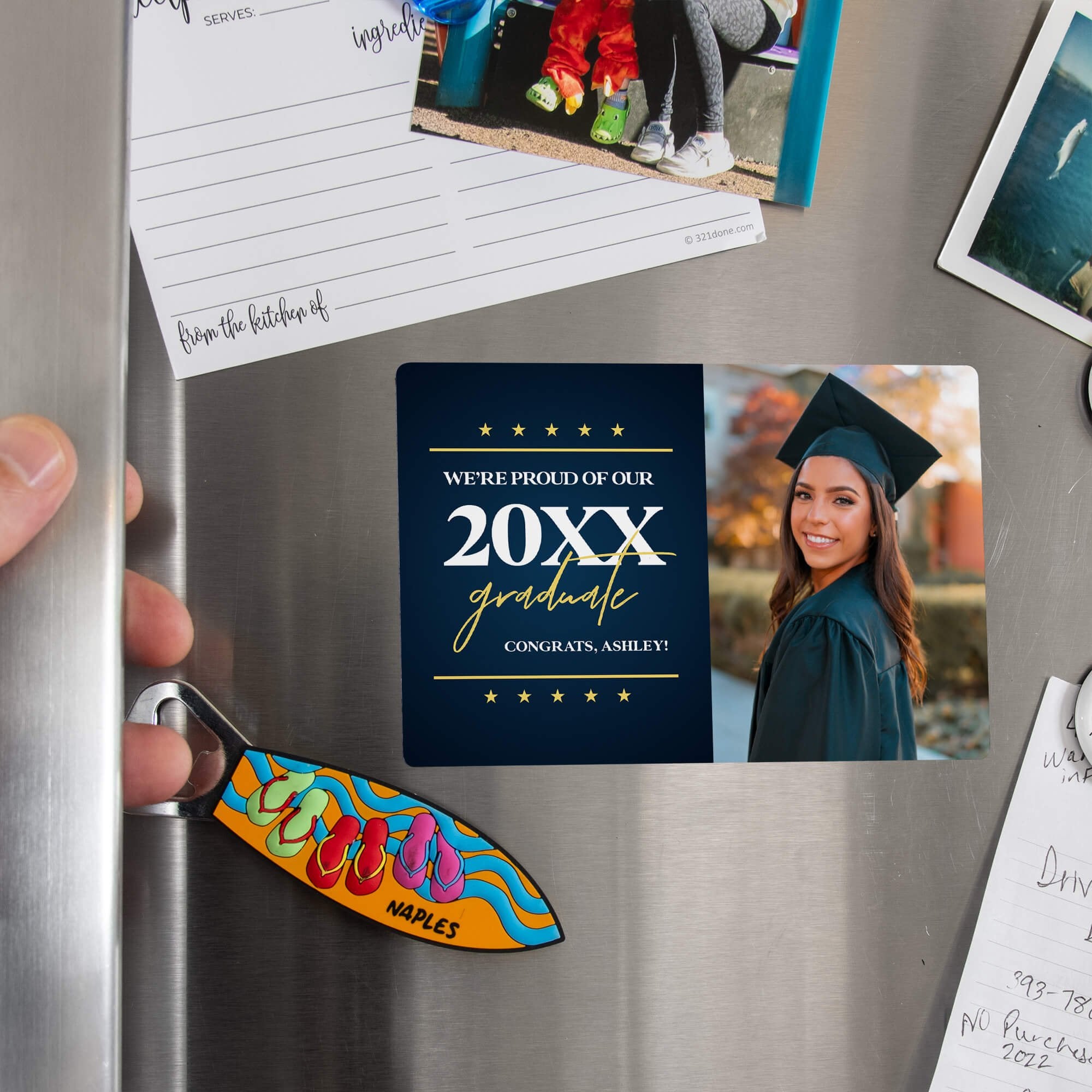 Hand placing a custom Star & Script Proud of Our Grad Graduation Announcement magnet on a stainless steel refrigerator