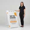 Woman standing beside a orange now hiring A-frame sign for scale