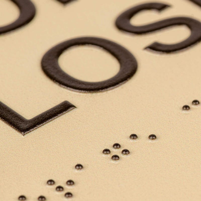 A close-up of the tactile letters and braille on the two-tone layered ADA sign