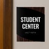 A modern twist design ADA sign for a student center with the dark wood background and black foreground