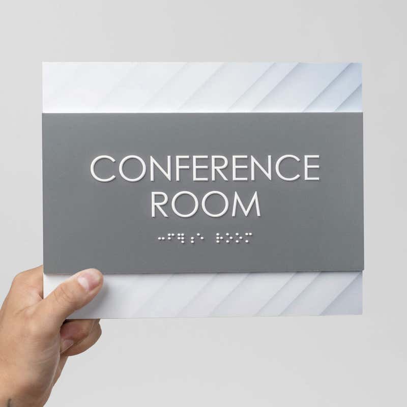 A person holding a 2-layered ADA sign for a conference room with a gray foreground and minimal diagonal background