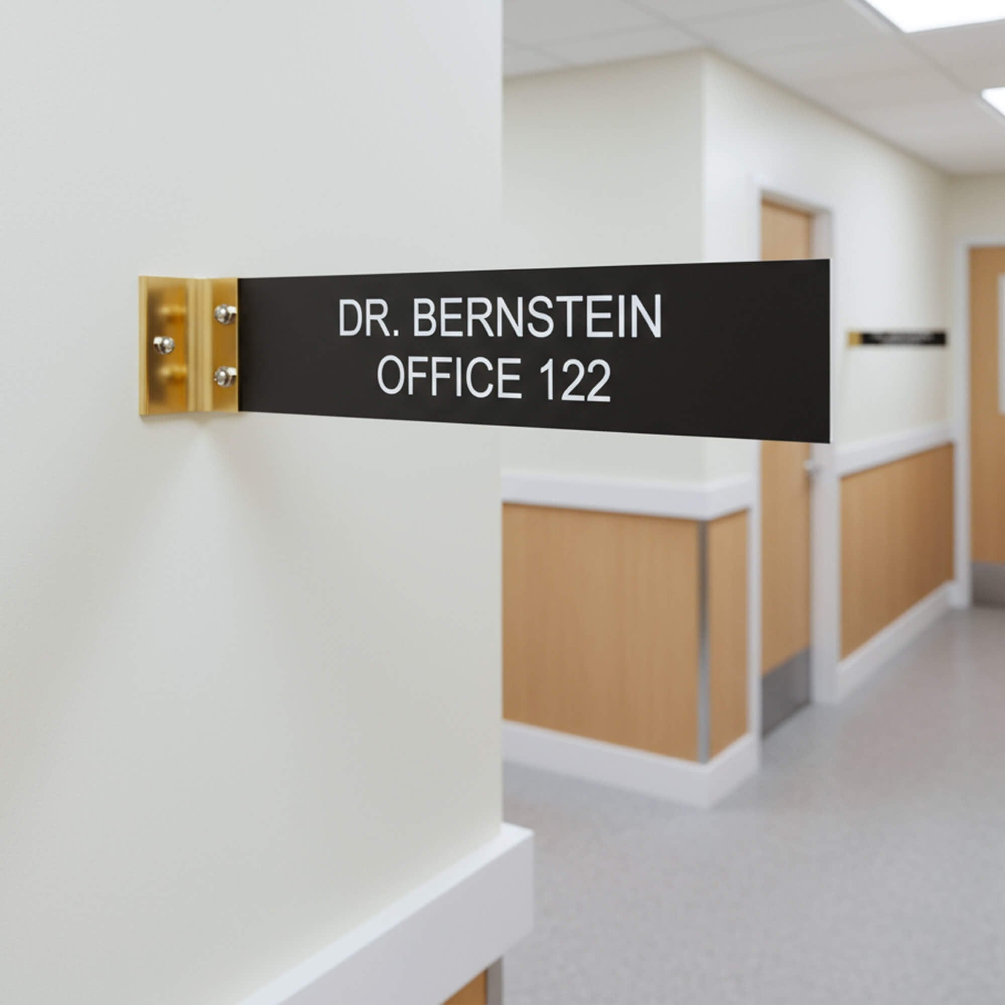 2” x 10” Corridor sign with black material color, white engraved text, and gold holder on a hallway wall with custom text saying “DR. BERNSTEIN OFFICE 122”.