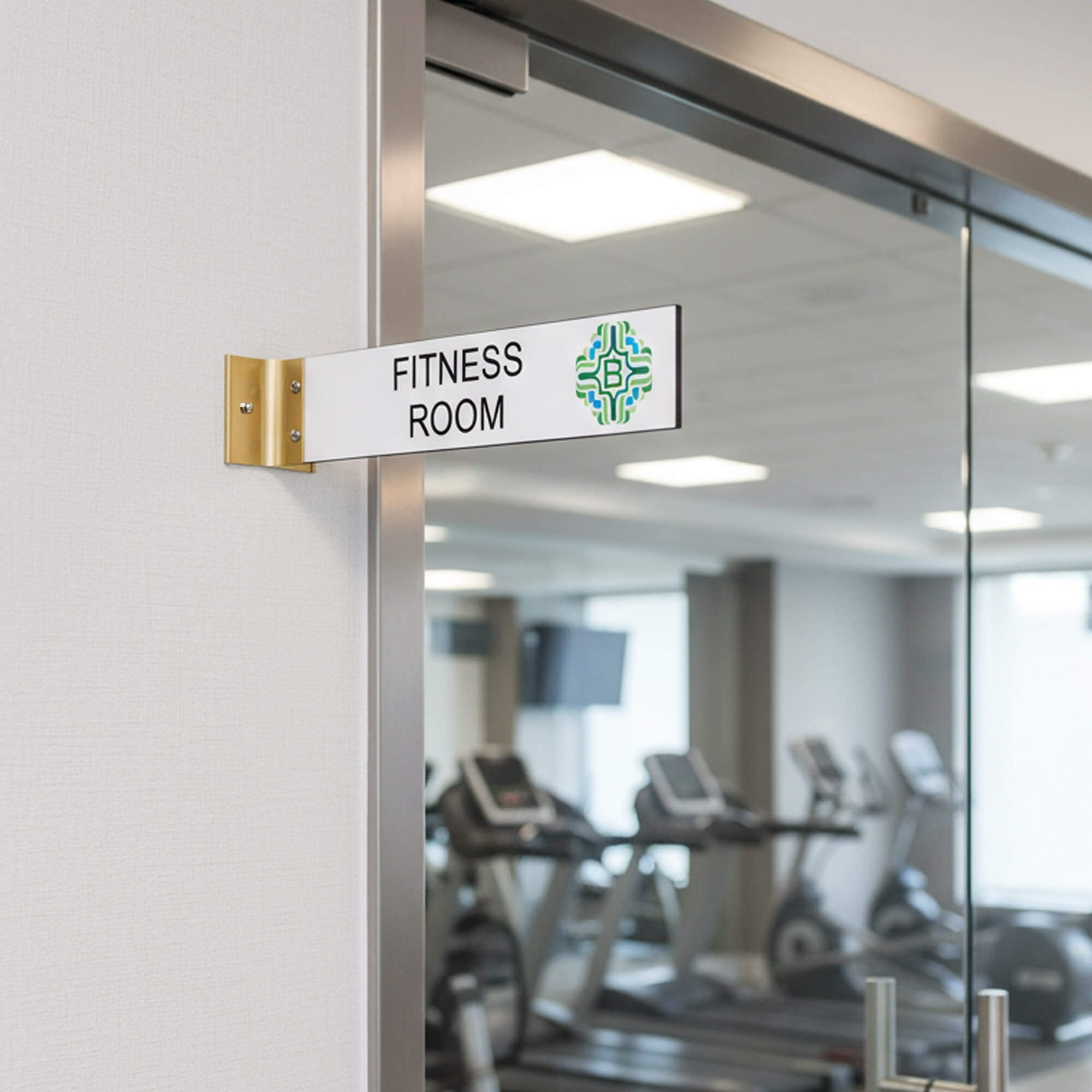2” x 10” Corridor sign with white material color, black engraved text, logo, and gold holder on a hallway wall with custom text saying “FITNESS ROOM”.
