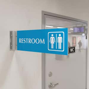 4” x 12” Corridor sign with baby blue material color, white engraved text, logo and silver holder on a hallway wall with text saying “RESTROOM”.