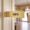4” x 12” Corridor sign with gold material color, black engraved text, and gold holder on a hallway wall with text saying “MEN”.