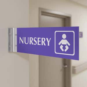 4” x 12” Corridor sign with purple material color, white engraved text, logo and silver holder on a hallway wall with text saying “NURSERY”.