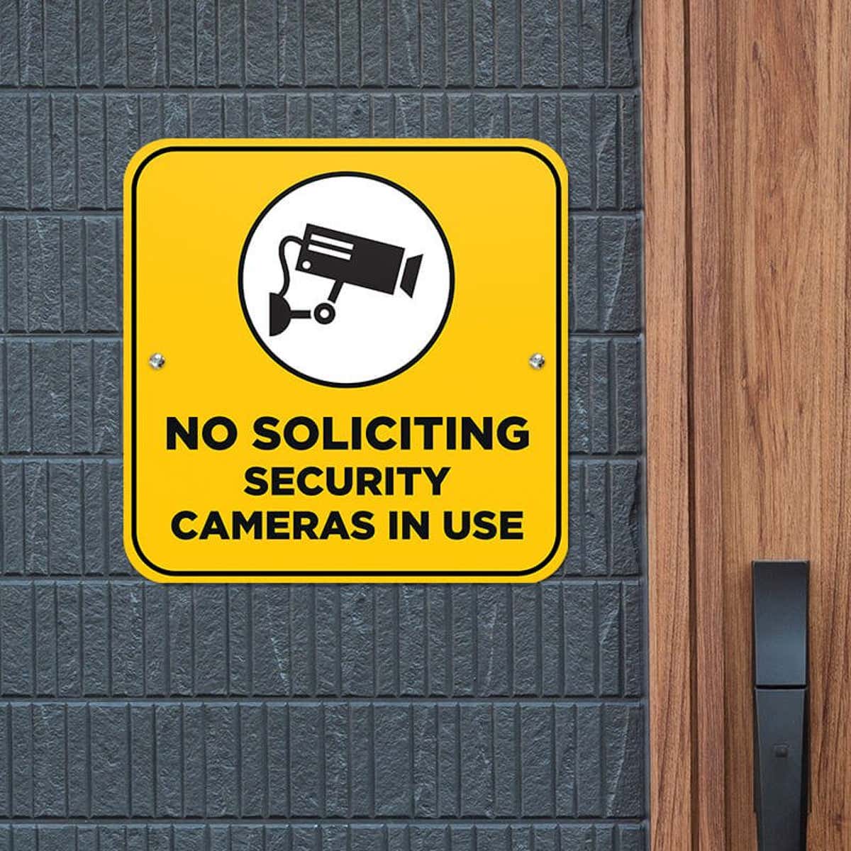 Yellow No Soliciting sign on a gray brick wall reading "Security Cameras in Use" with a security camera icon