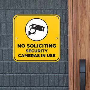 Yellow No Soliciting sign on a gray brick wall reading "Security Cameras in Use" with a security camera icon