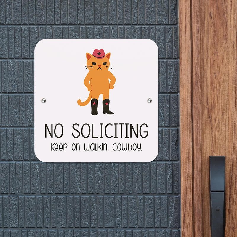 Cute orange frowning cat no soliciting sign that reads, “Keep on walkin, cowboy” hung up on wall