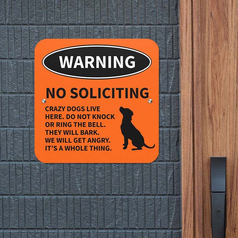 No soliciting dog warning sign with dog icon with bold text reading: “Crazy dogs live here” hung up on a gray wall