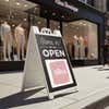 A sidewalk sign displayed outside of a boutique that says We Are Open and features their logo