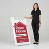 A woman standing next to an open house A-frame sign with her hand on the top of the sign